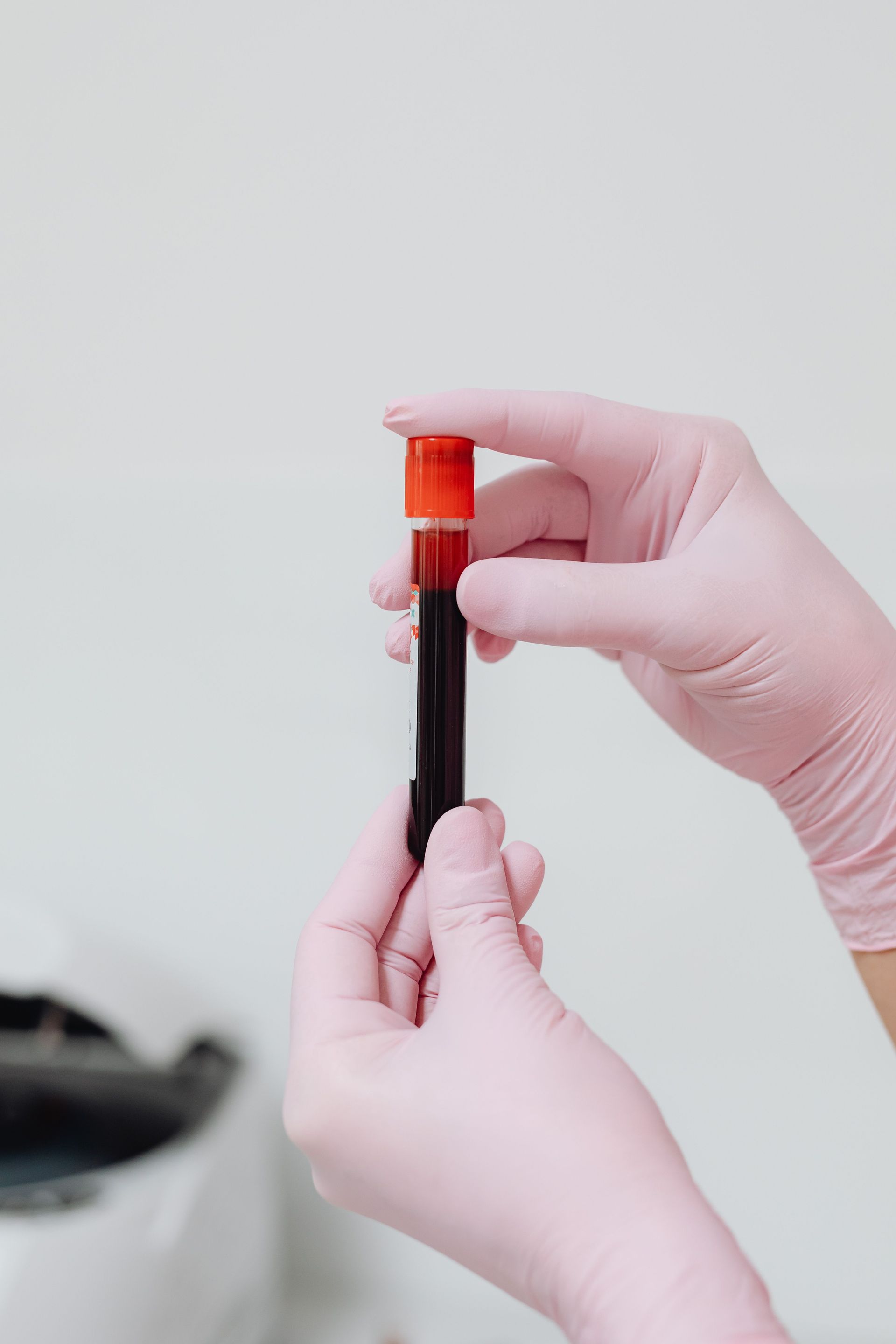 Close-up of gloved hands holding a blood test tube with red liquid against a white background.